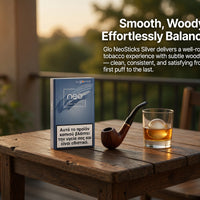 A pack of Glo NeoSticks Silver tobacco pods stands on a rustic wooden table beside a smoking pipe and a glass of amber liquid, with outdoor patio furniture and warm sunset light in the background. White promotional text appears on the right side.
