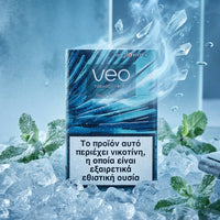 A pack of Veo Tobacco Products disposable cigarettes stands upright in a bed of ice with mint leaves and a vape device nearby. The packaging is blue and black with a large white health warning in Greek and the brand name 'veo' prominently displayed.