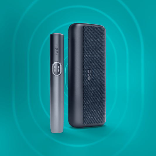 The image shows two components of the IQOS ILUMA i PRIME Kit against a turquoise background. On the left is a slim, silver-colored device with the IQOS logo, and on the right is a larger, black textured device.