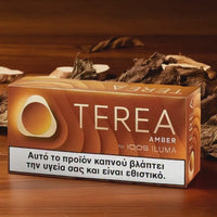 A box of Terea Amber tobacco by IQOS ILUMA on a wooden surface with dried leaves and wood chips. The packaging is brown with a white warning label at the bottom.