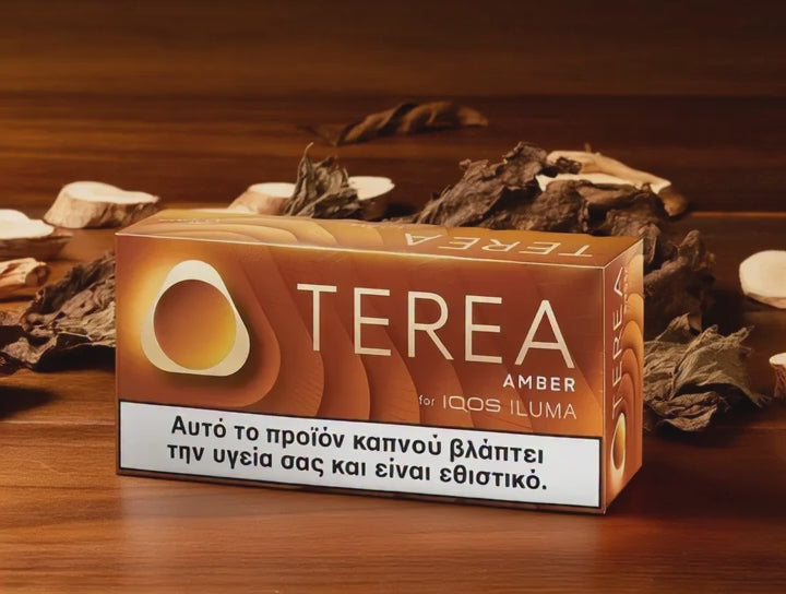 A box of Terea Amber tobacco by IQOS ILUMA on a wooden surface with dried leaves and wood chips. The packaging is brown with a white warning label at the bottom.