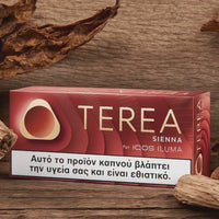 A box of OTEREA Sienna for IQOS ILUMA cigarettes placed on a rustic wooden surface, surrounded by dried tobacco leaves. The packaging is dark red with gold lettering and a white health warning label at the bottom.