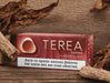 A box of OTEREA Sienna for IQOS ILUMA cigarettes placed on a rustic wooden surface, surrounded by dried tobacco leaves. The packaging is dark red with gold lettering and a white health warning label at the bottom.