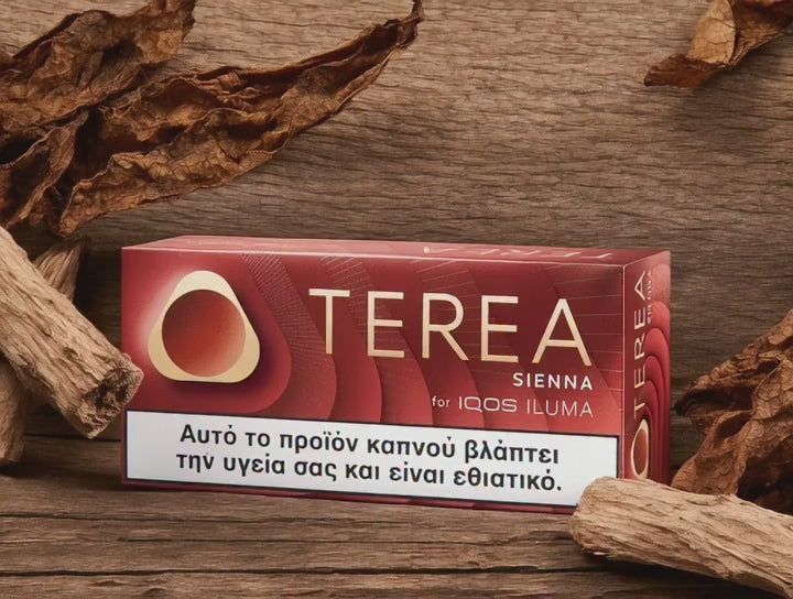 A box of OTEREA Sienna for IQOS ILUMA cigarettes placed on a rustic wooden surface, surrounded by dried tobacco leaves. The packaging is dark red with gold lettering and a white health warning label at the bottom.