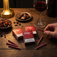 A hand holds a red EVO disposable vape pen on a rustic wooden table, with three red EVO product boxes, several loose pens, a glass of red wine, chocolate pieces, and dried fruit in the background.