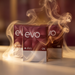 Three packs of EVO disposable cigarettes by Ploom Club arranged on a tabletop with dramatic warm lighting and swirling smoke effects. The front pack is fully visible, with two partially obscured packs behind it.
