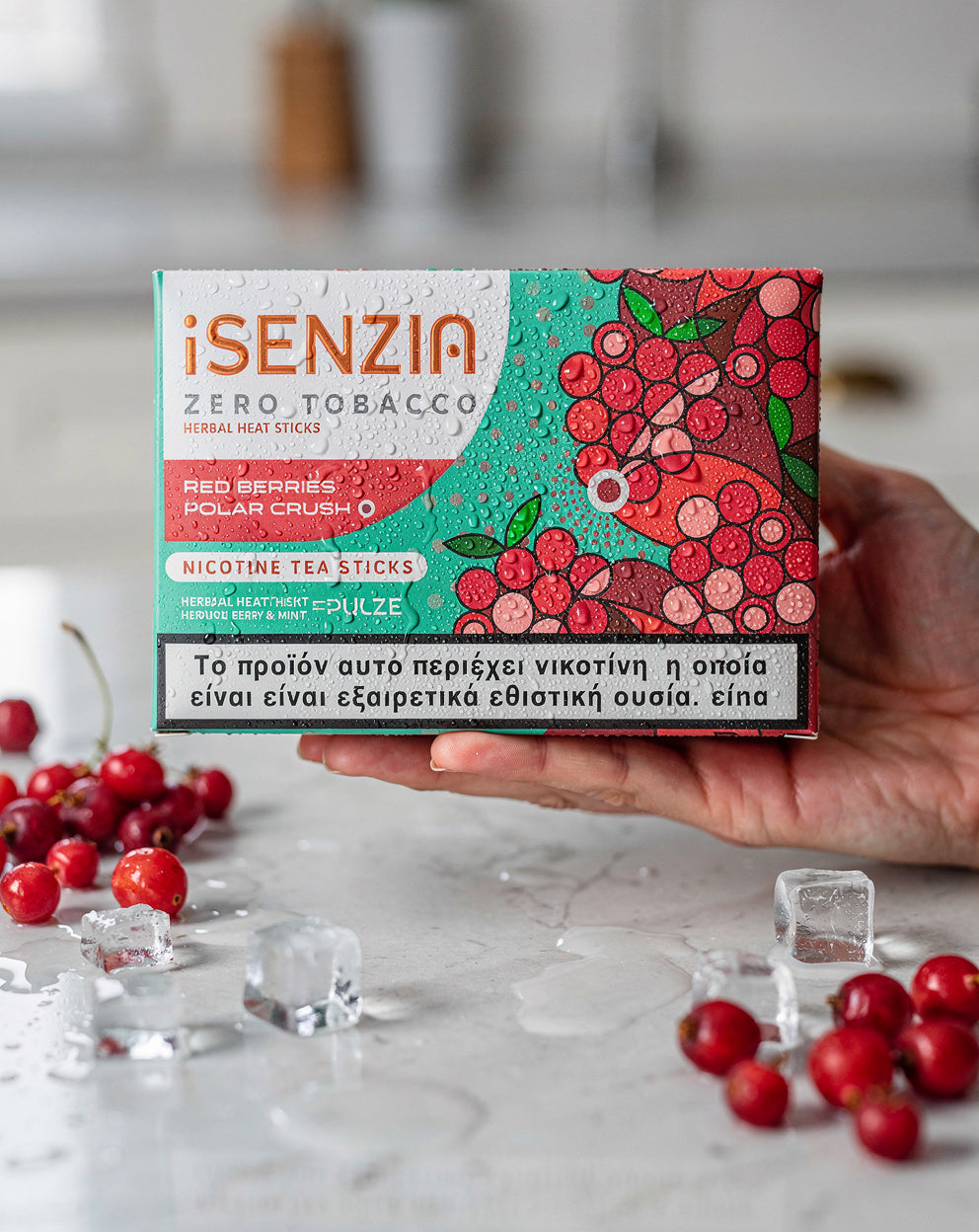 A hand holds a box of iSENZIA Zero Tobacco Red Berries Polar Crush nicotine tea sticks. The packaging features a vibrant red and green berry illustration, with scattered red berries and ice cubes on a light marble surface in the foreground.